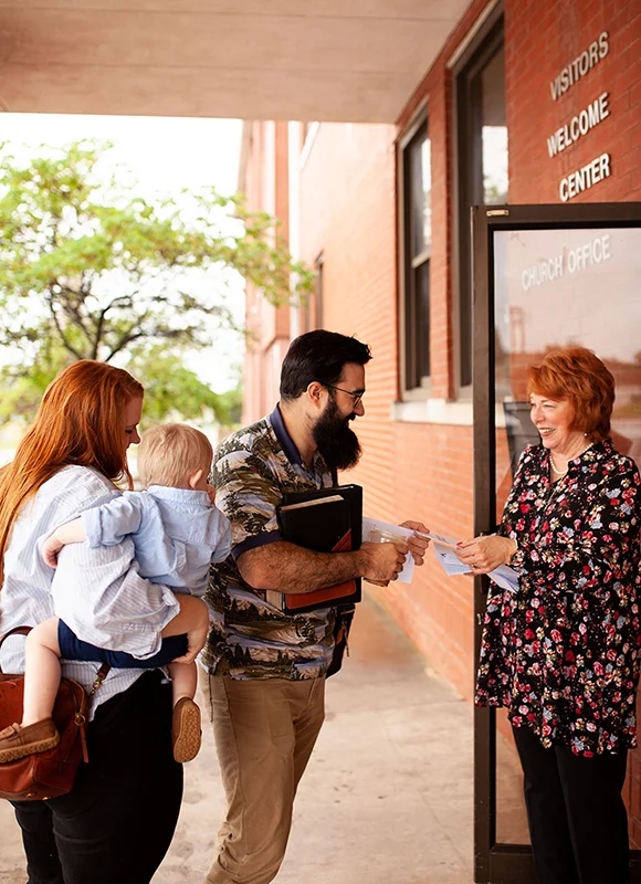 A young family with a child being warmly greeted by a woman handing out bulletins at the entrance of a church building, creating a welcoming atmosphere.