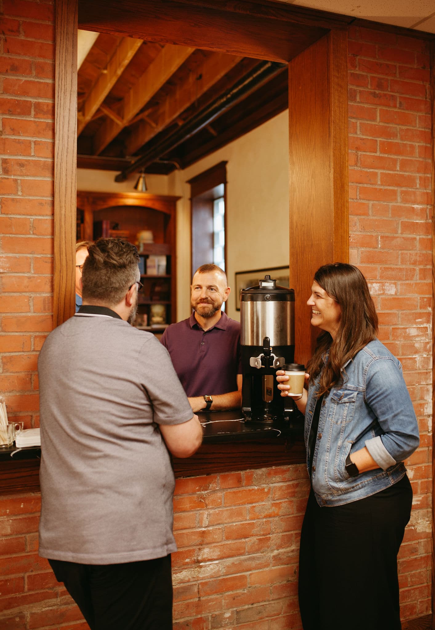 A group of individuals enjoying coffee and conversation in a warm, rustic community space with exposed brick walls and wooden accents.