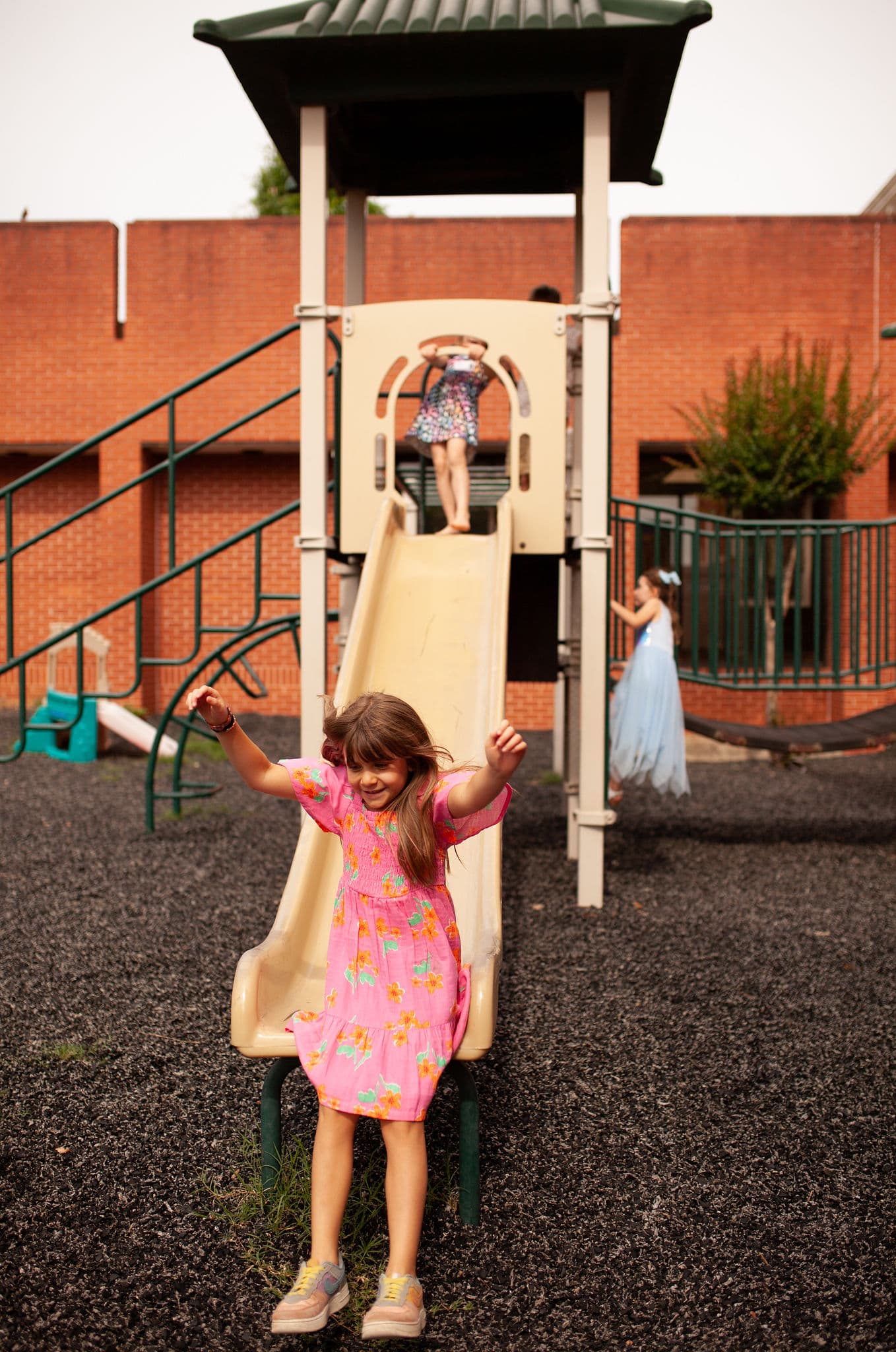 A young girl in a pink dress happily sliding down a playground slide, with other children playing in the background on a sunny day.